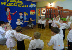 Odświętnie ubrane dzieci tańczą w kole na tle dekoracji. Dwie nauczycielki tańczą z dziećmi.
